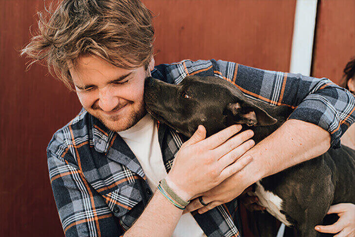 Mann, der einen schwarzen Hund liebevoll umarmt, beide sehen glücklich aus und stehen sich nahe.