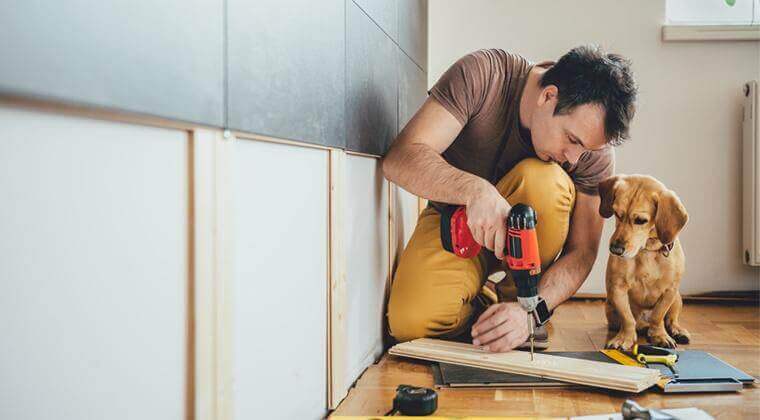Un homme utilise une perceuse pendant qu'un chien curieux observe attentivement.
